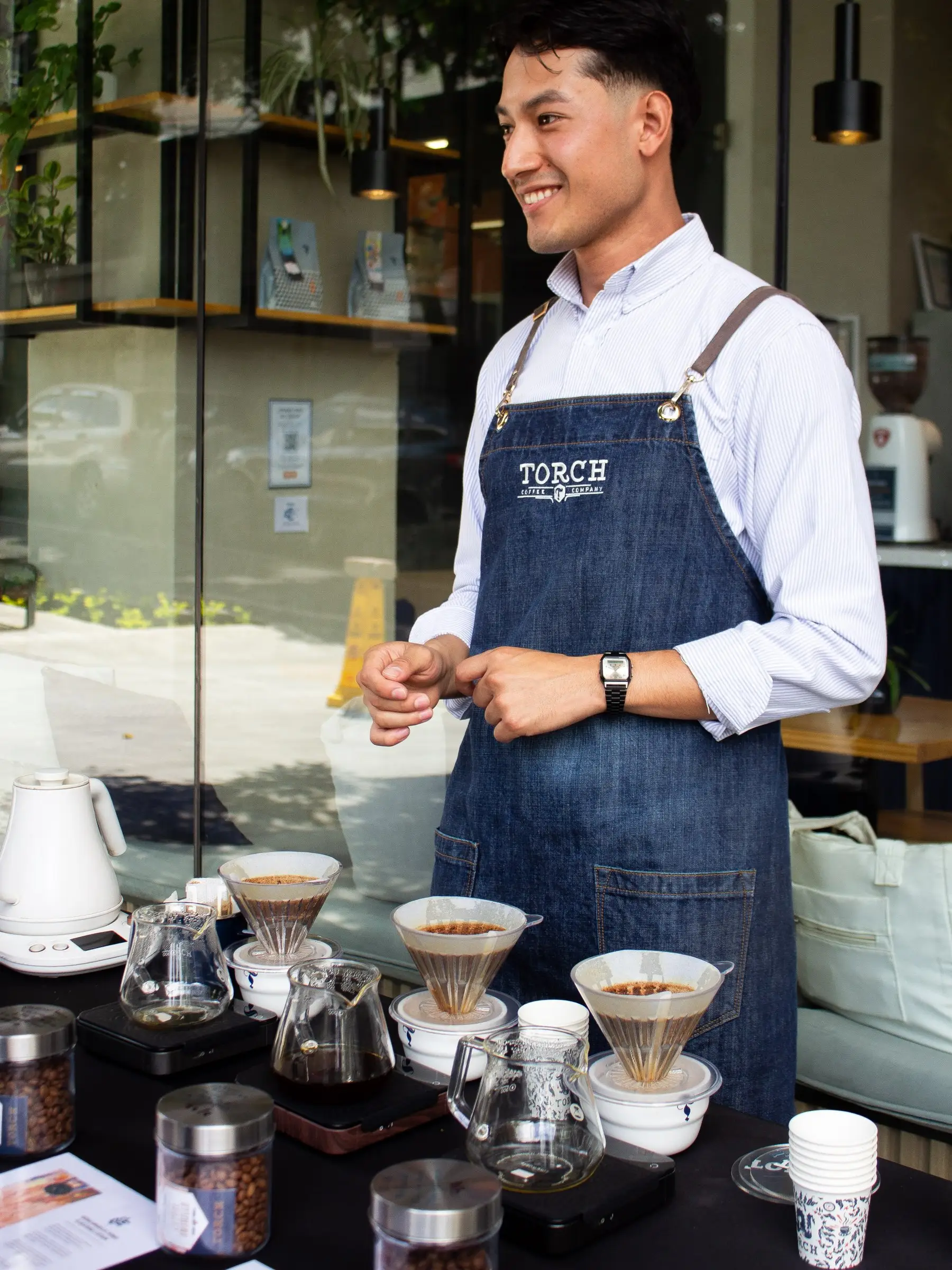 Barista de Torch sonriendo frente a una mesa de métodos de filtrado en la entrada de la sede de Zona 4